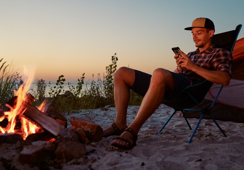 man on camping trip sitting in front of fire with cell phone on hand